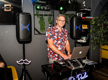 DJ Radu bag pladerne i blomstret skjorte, ved siden af højtalere og neonskiltet 'DJ Radu'