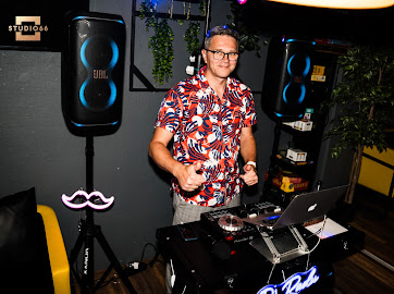 DJ Radu smiler bag pladerne, ved siden af højtalere og neonskiltet 'DJ Radu'