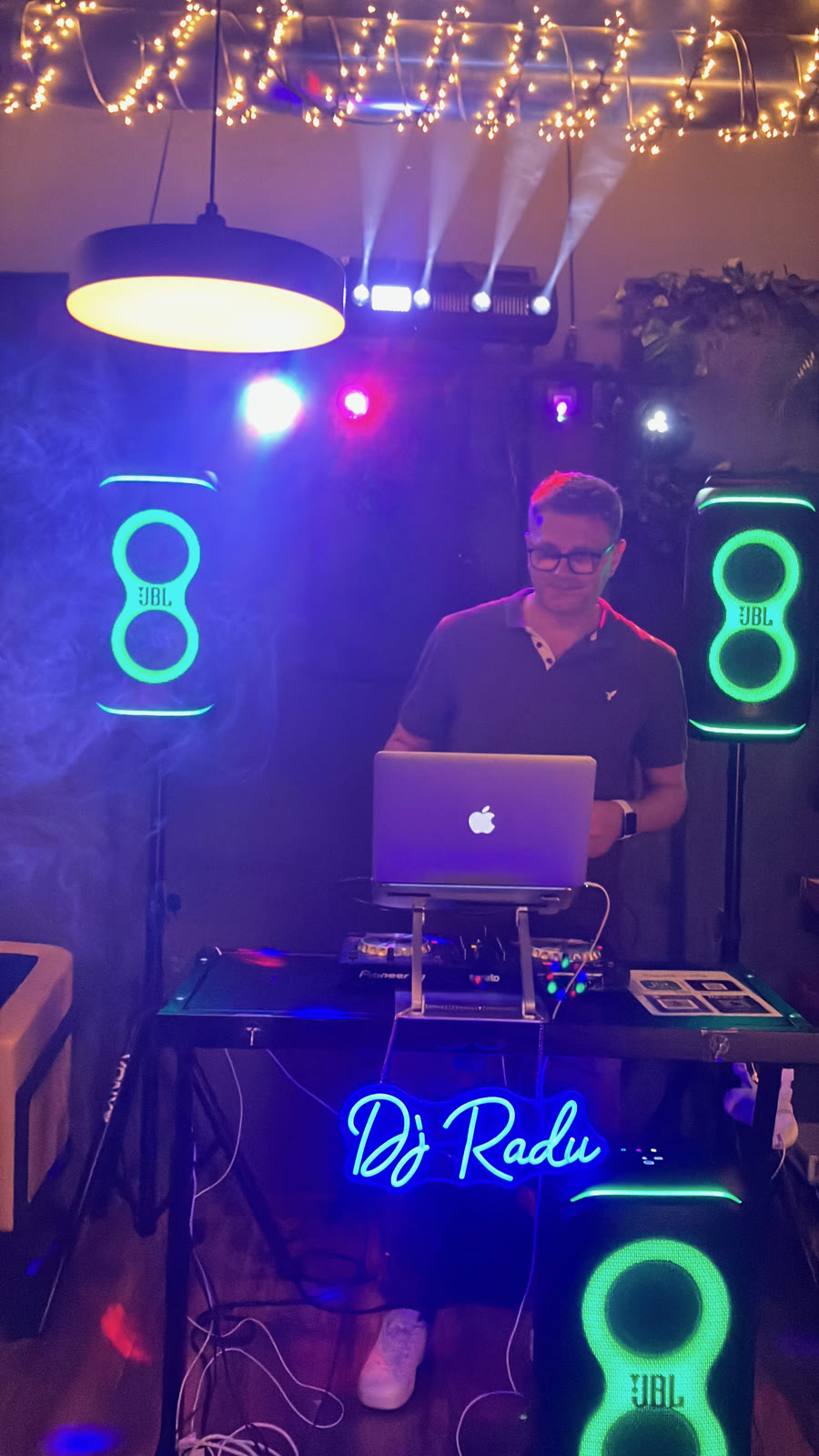 DJ Radu la pupitru, sub semnul neon „DJ Radu” și ghirlandele de lumini